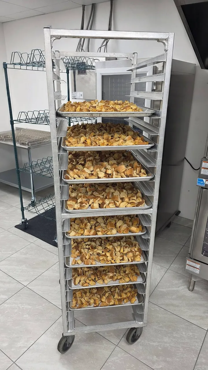A tall rolling rack lined with trays of bread cubes drying for bread pudding